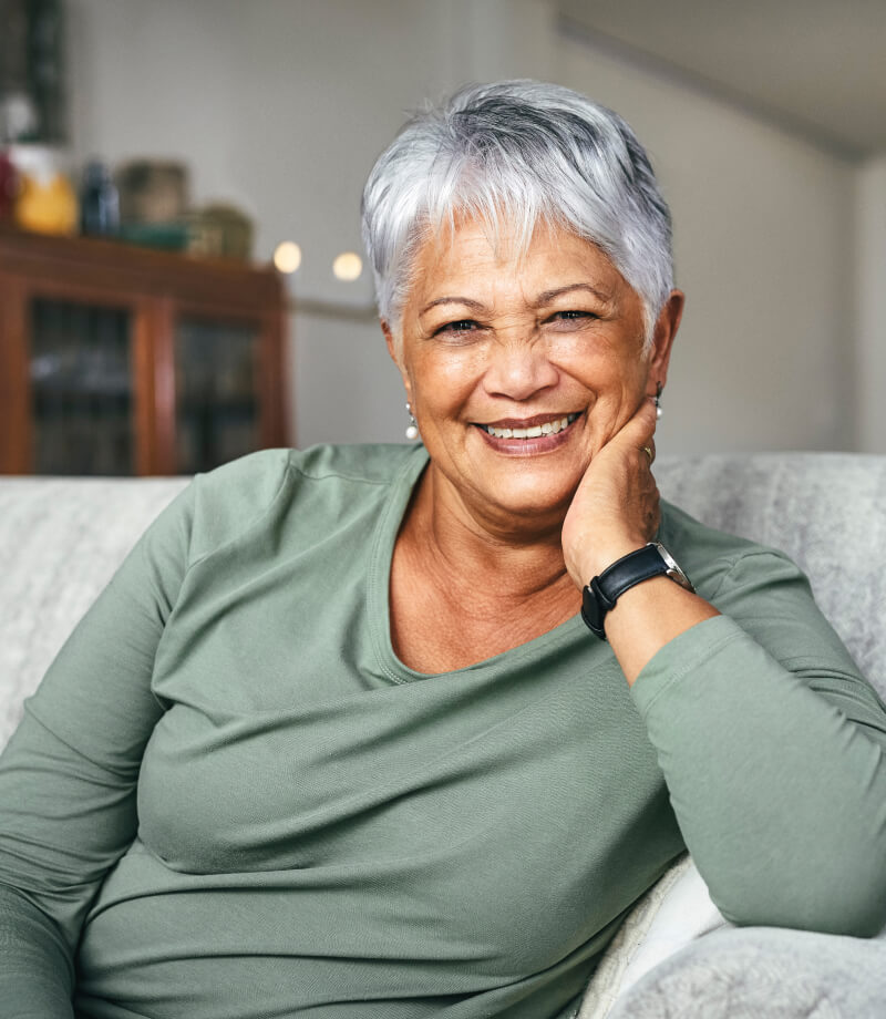 Smiling woman with short gray hair sitting on a couch, resting her head on her hand.