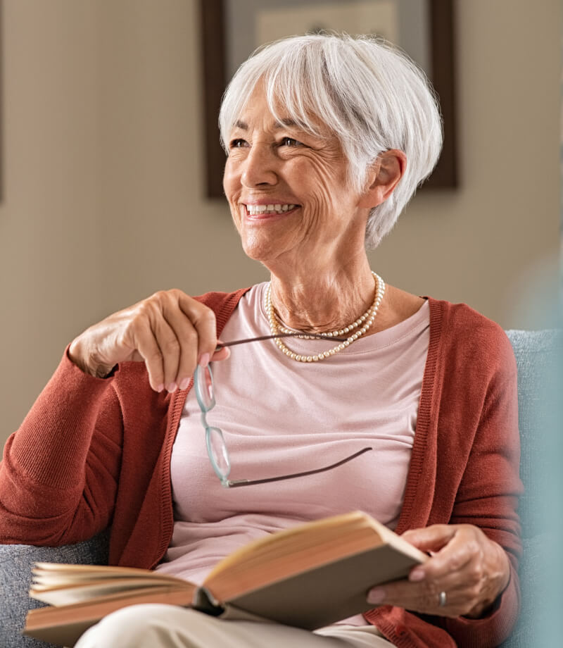 Smiling woman with short gray hair holding glasses and a book while seated indoors.