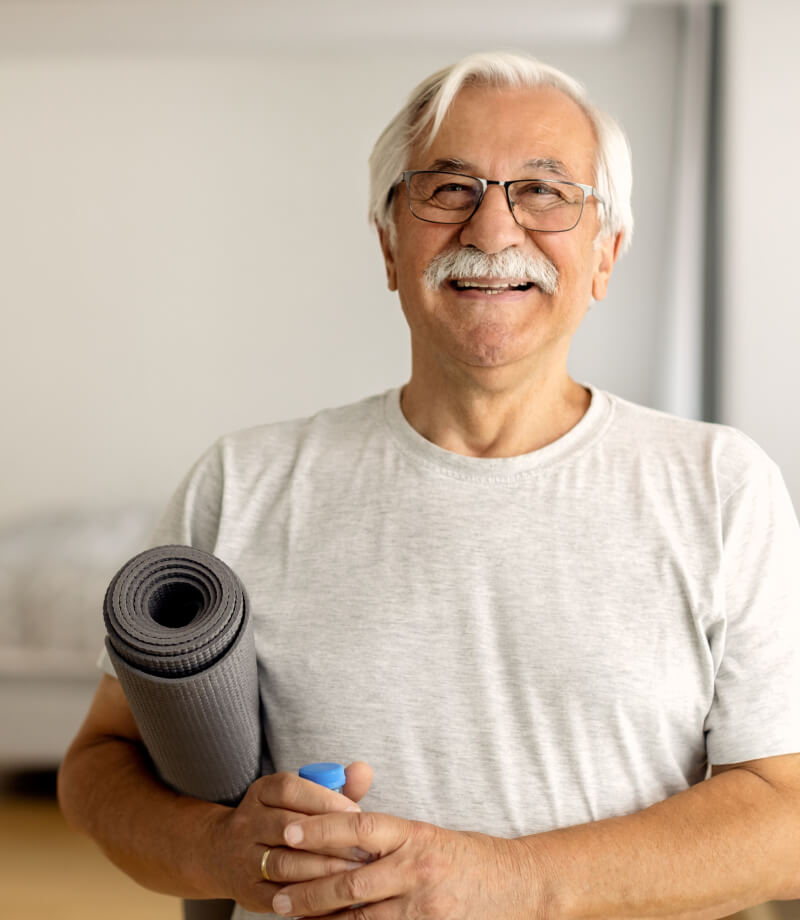Smiling older man with glasses holding a rolled-up yoga mat and water bottle.
