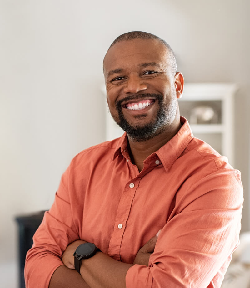 Smiling man in an orange shirt standing with arms crossed indoors.